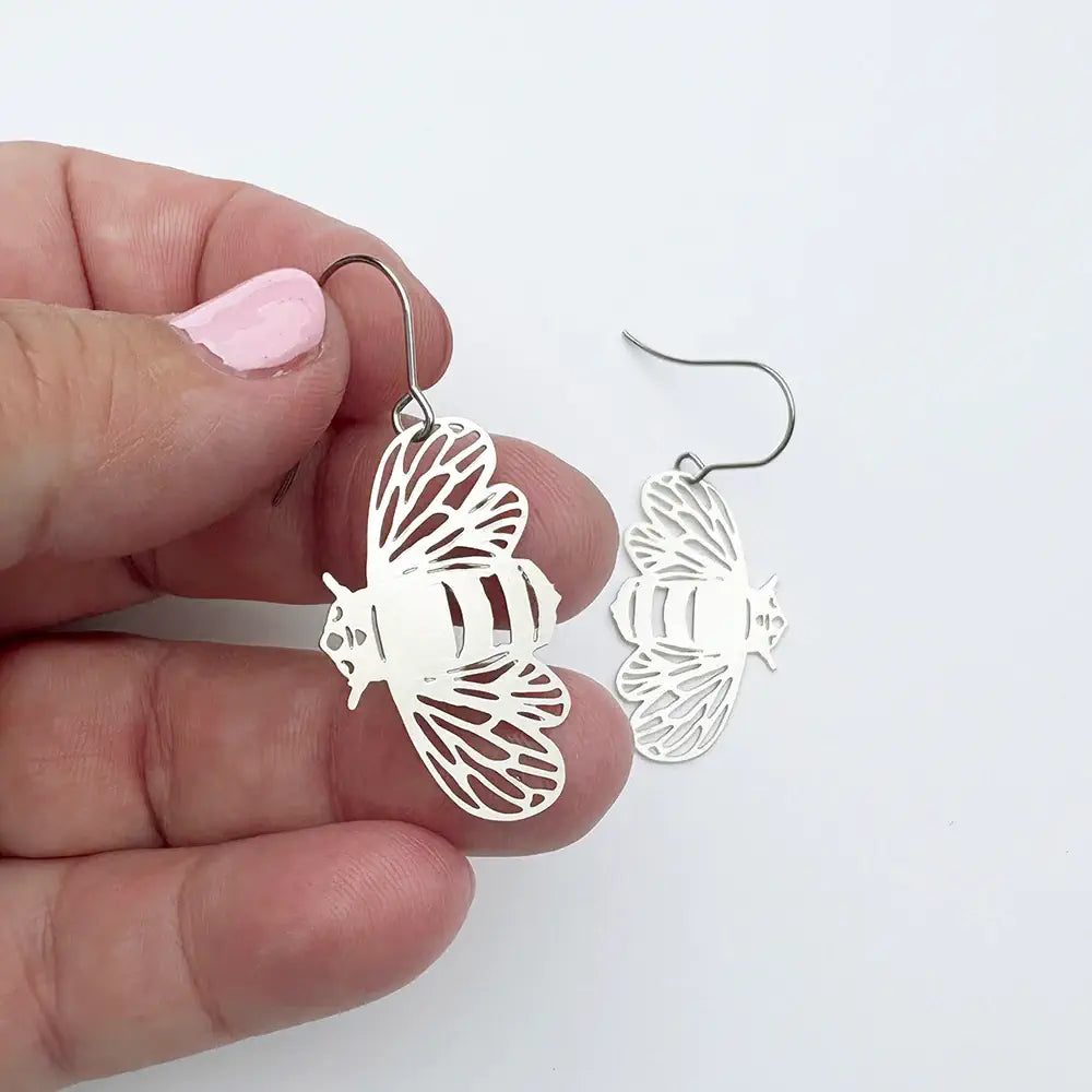 Silver bee-shaped earrings held by a hand with pink nail polish on a white background