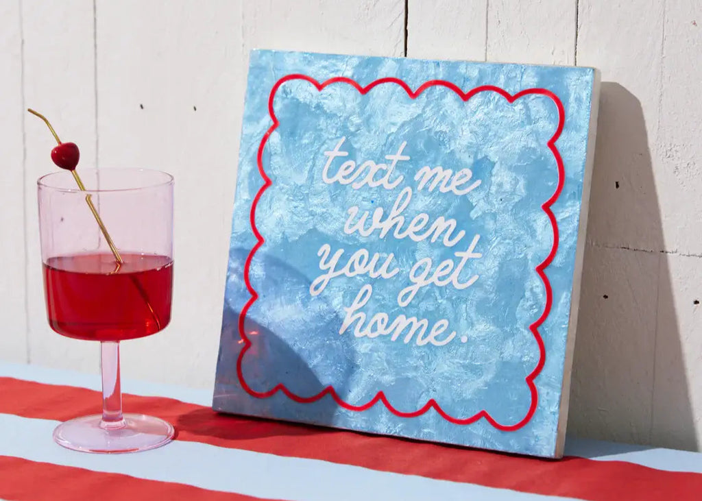 Canvas with 'text me when you get home' quote on a red and white striped surface with a drink.