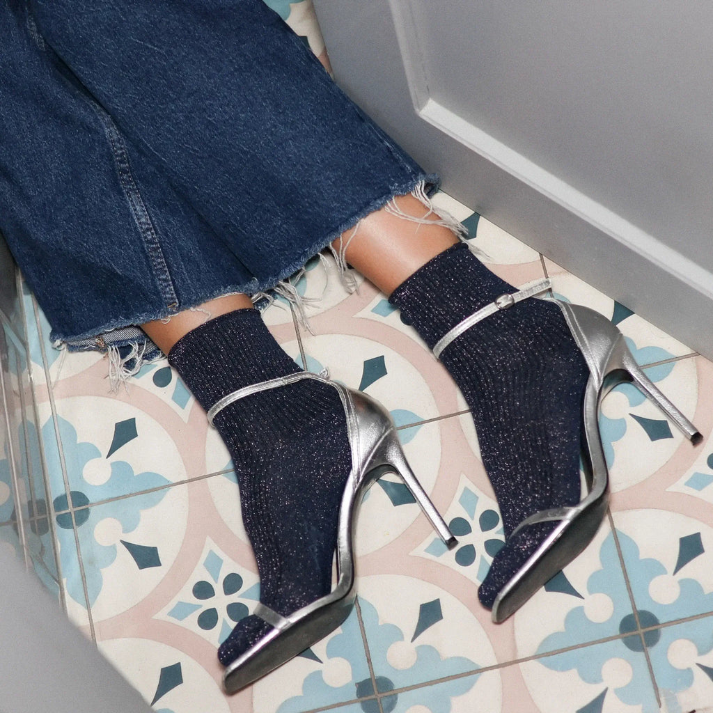 Blue glittery high-heeled shoes worn with jeans on a patterned floor.