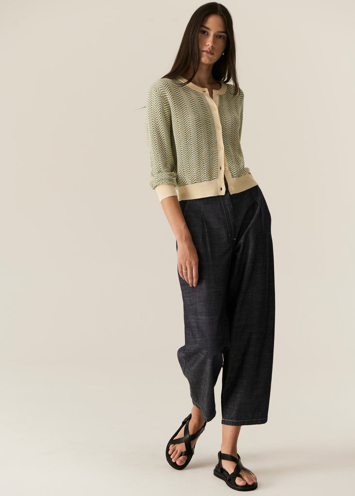 Woman wearing a green patterned cardigan and black pants on a plain background