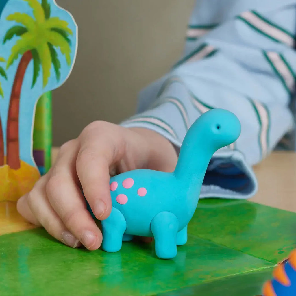 Child's hand holding a blue dinosaur toy on a green surface with a colorful card in the background