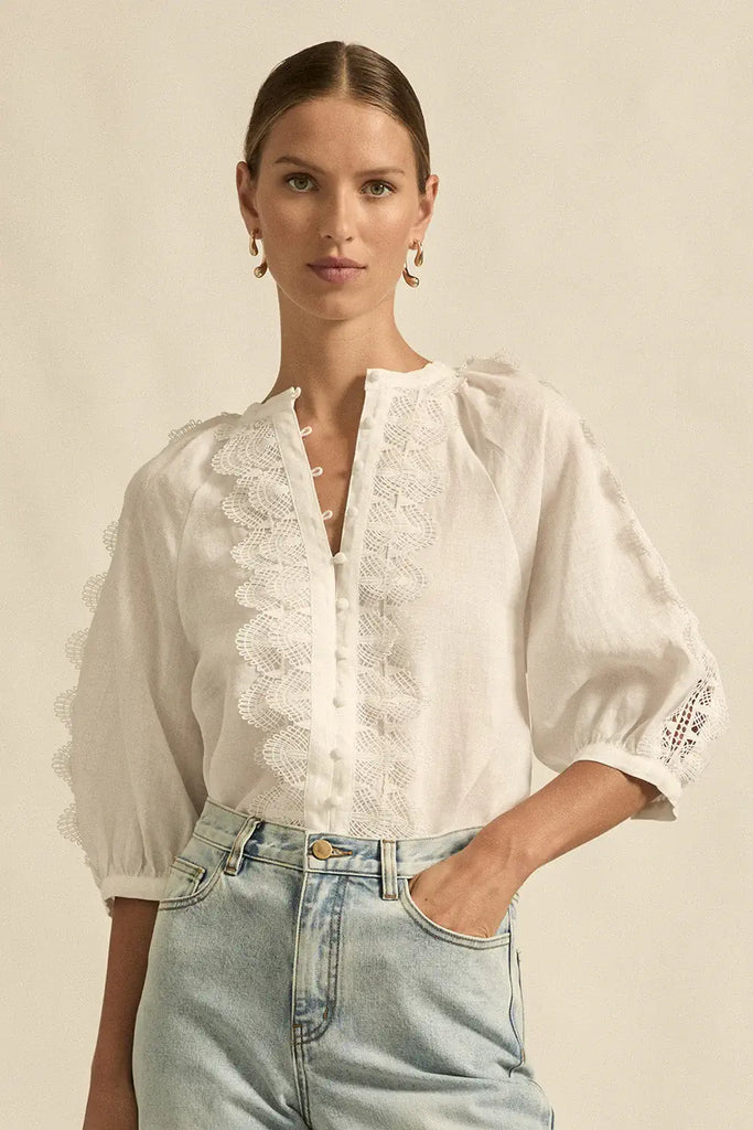 Woman wearing a white blouse with lace details and light blue jeans against a beige background