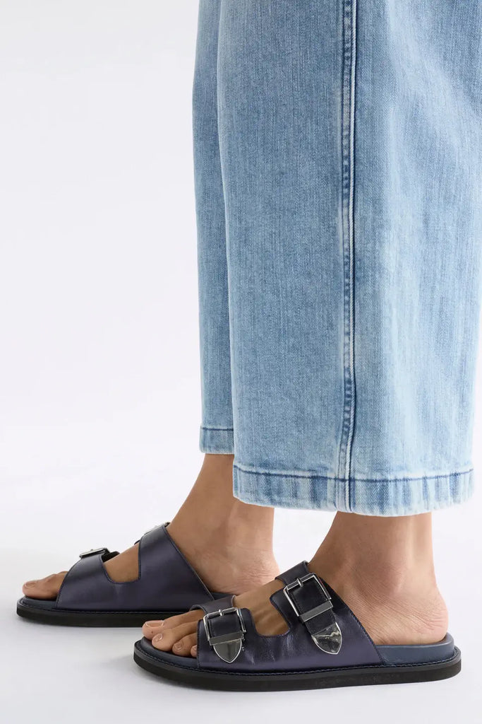 Navy sandals with buckles worn with light blue jeans on a white background