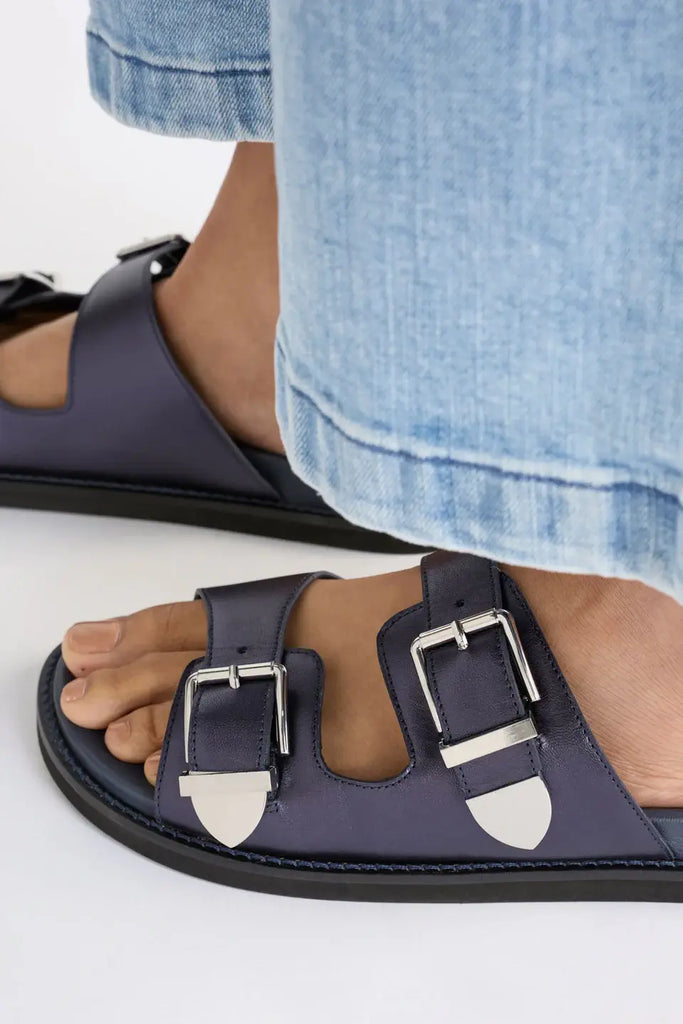 Navy sandals with buckles worn with light blue jeans on a white background