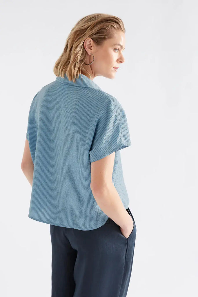 Woman wearing a light blue textured shirt and dark pants on a white background