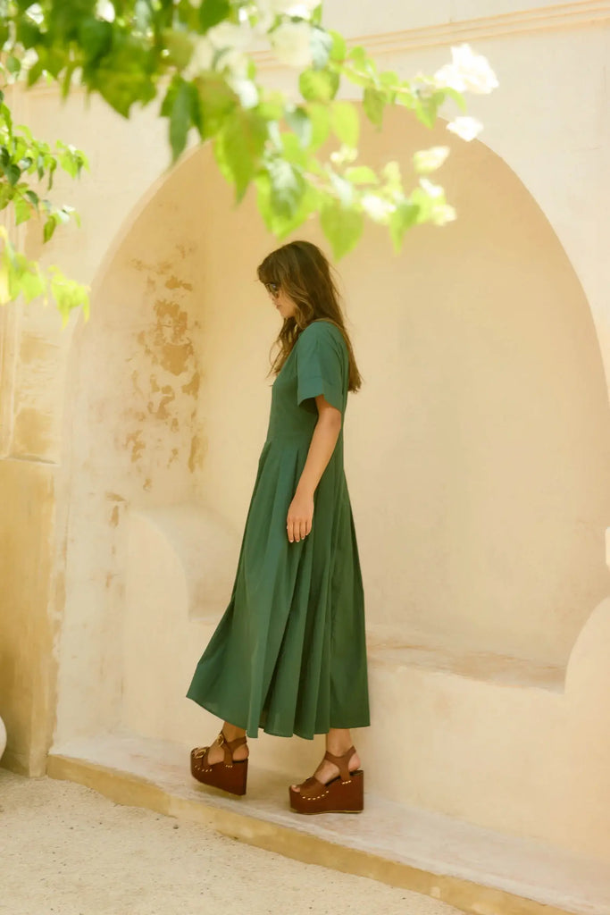 Woman in a green dress standing in a sunlit archway with greenery.
