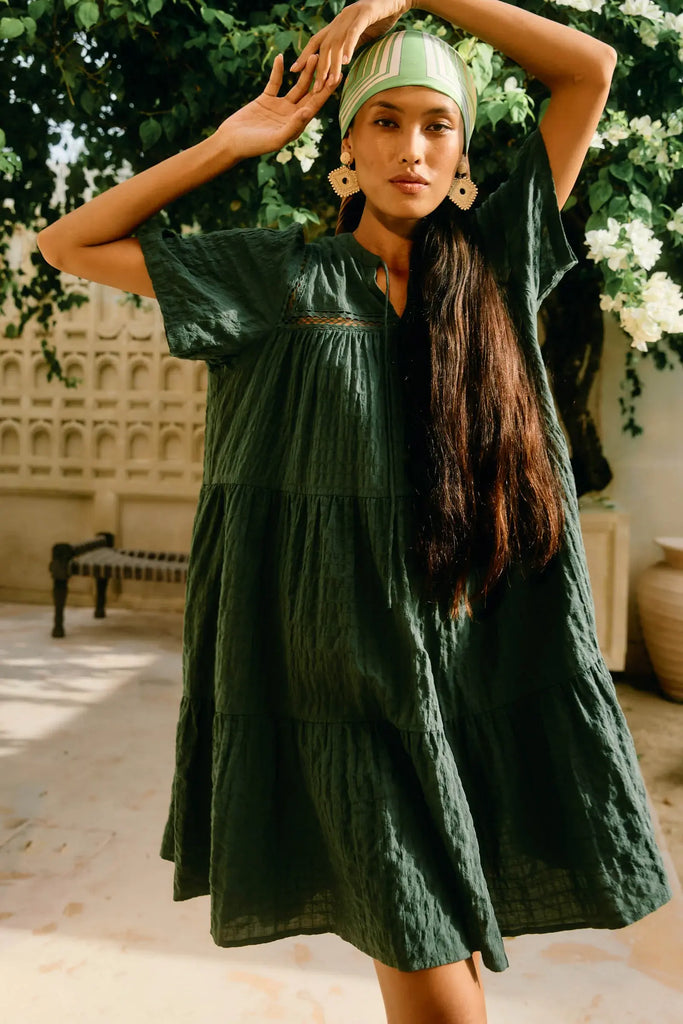 Woman wearing a green dress and headscarf in an outdoor setting with plants and a bench.