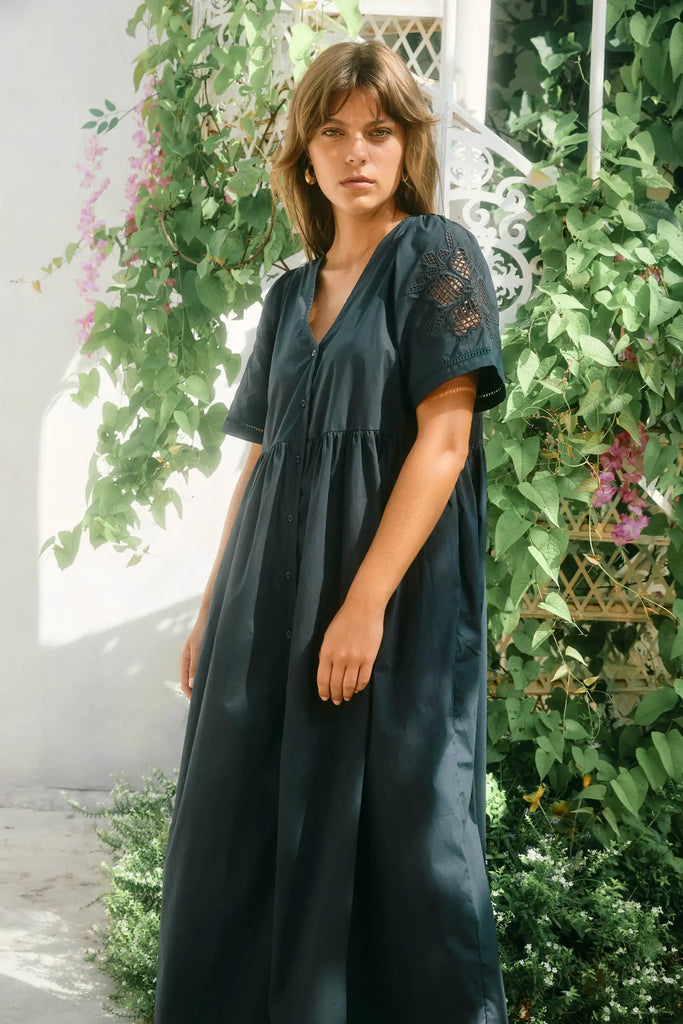 Woman wearing a dark green dress standing in a garden with greenery and flowers.
