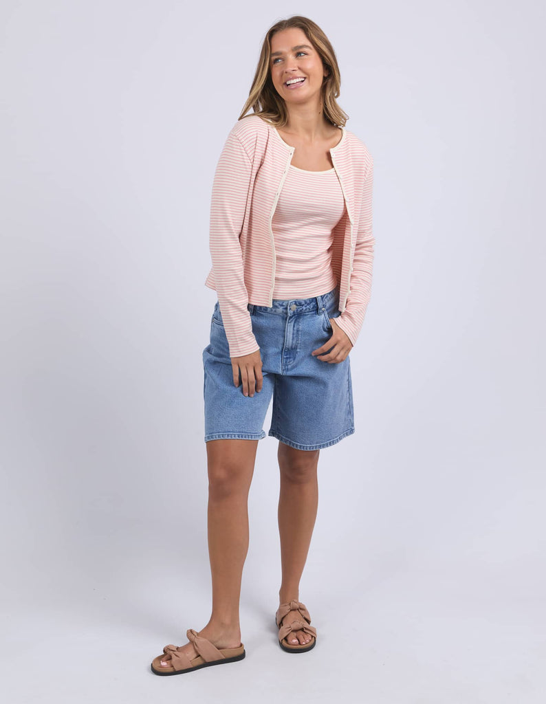 Woman wearing a pink cardigan, beige top, blue denim shorts, and sandals on a white background